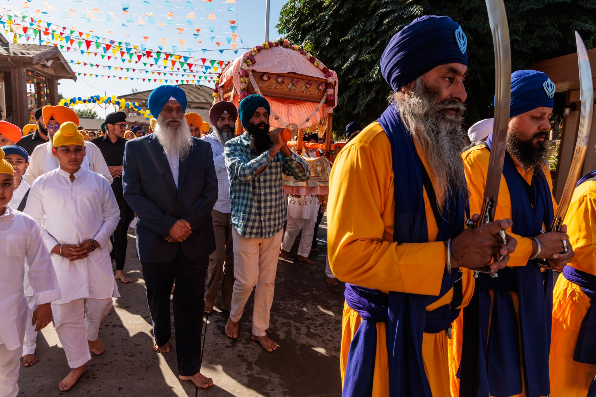 Two men with beards, wearing blue turbans and gold robes, hold swords as they walk in front of other people in turbans 