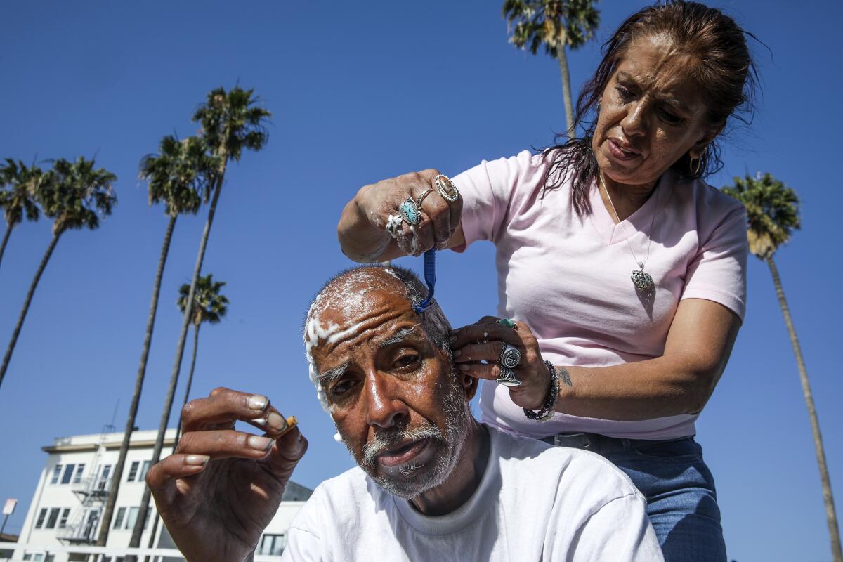 A seated gets his head shaved by a woman outdoors beneath palm trees.