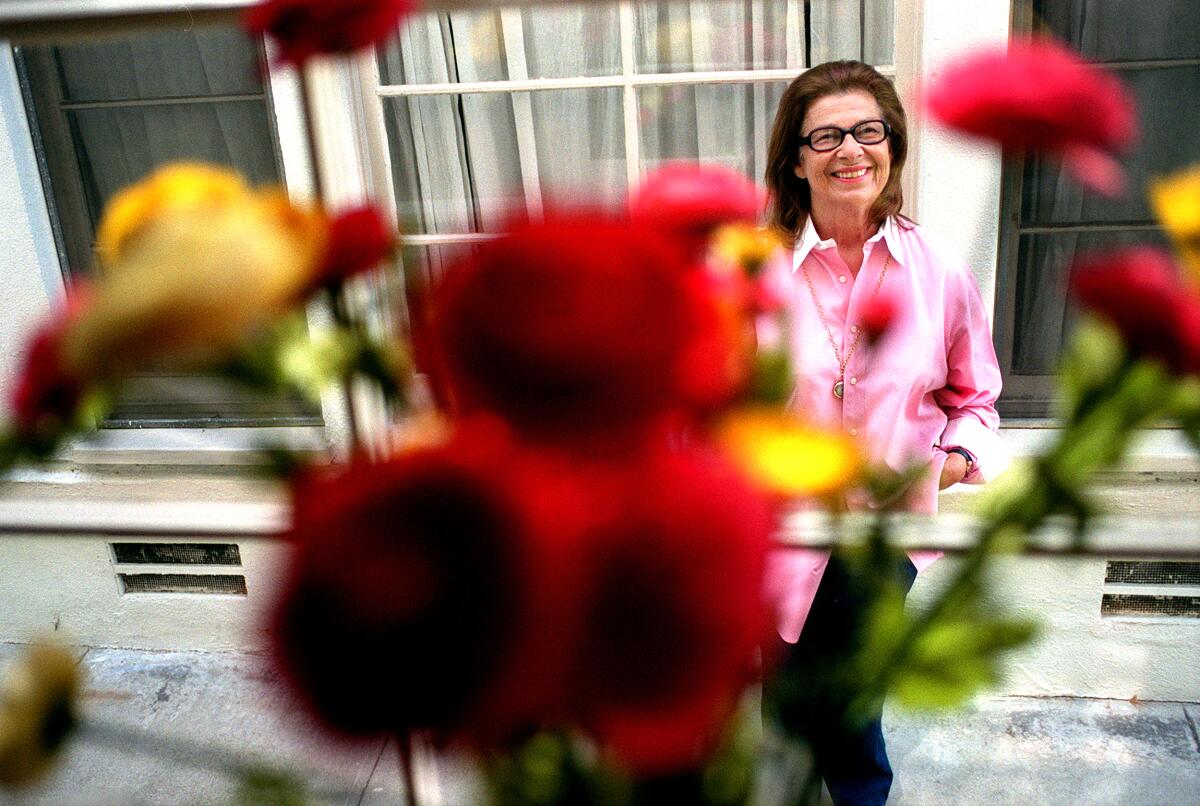Blurred flowers are in the foreground of a portrait of Norma Barzman, who smiles while sitting in a pink blouse