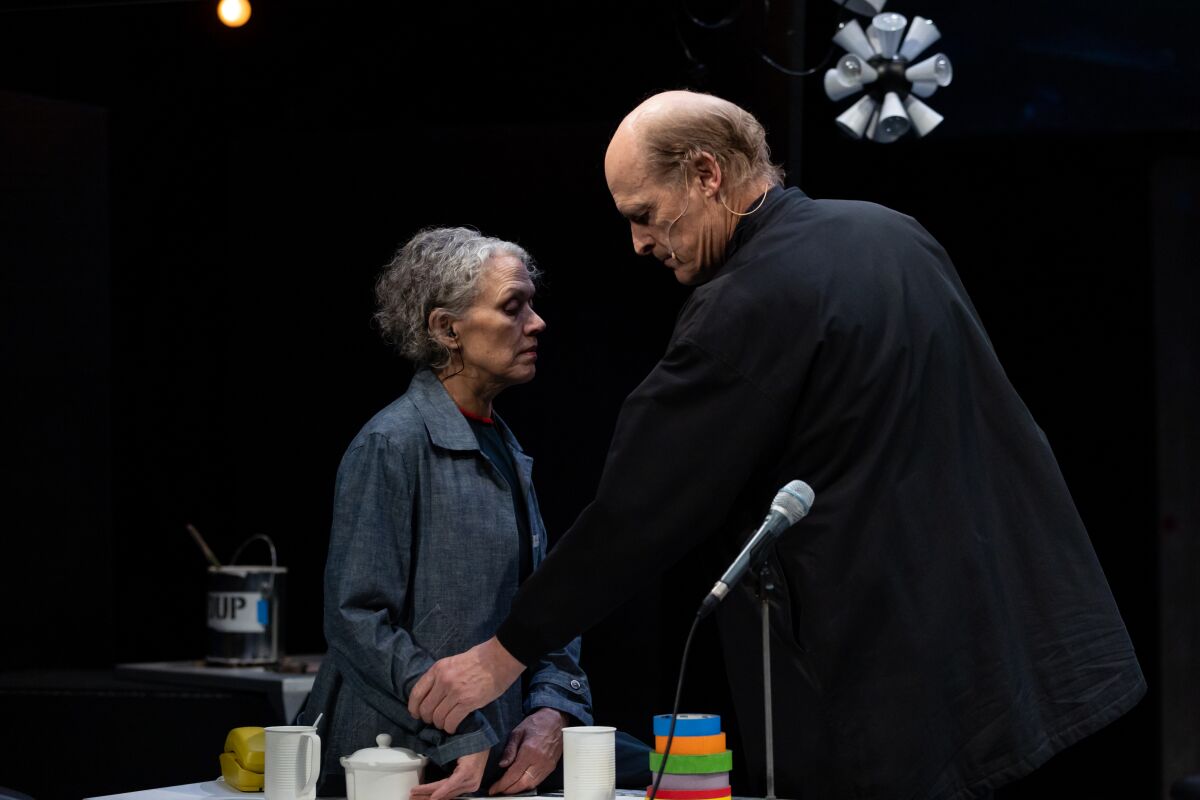 A woman and man face each other on a stage. He grasps her hand.