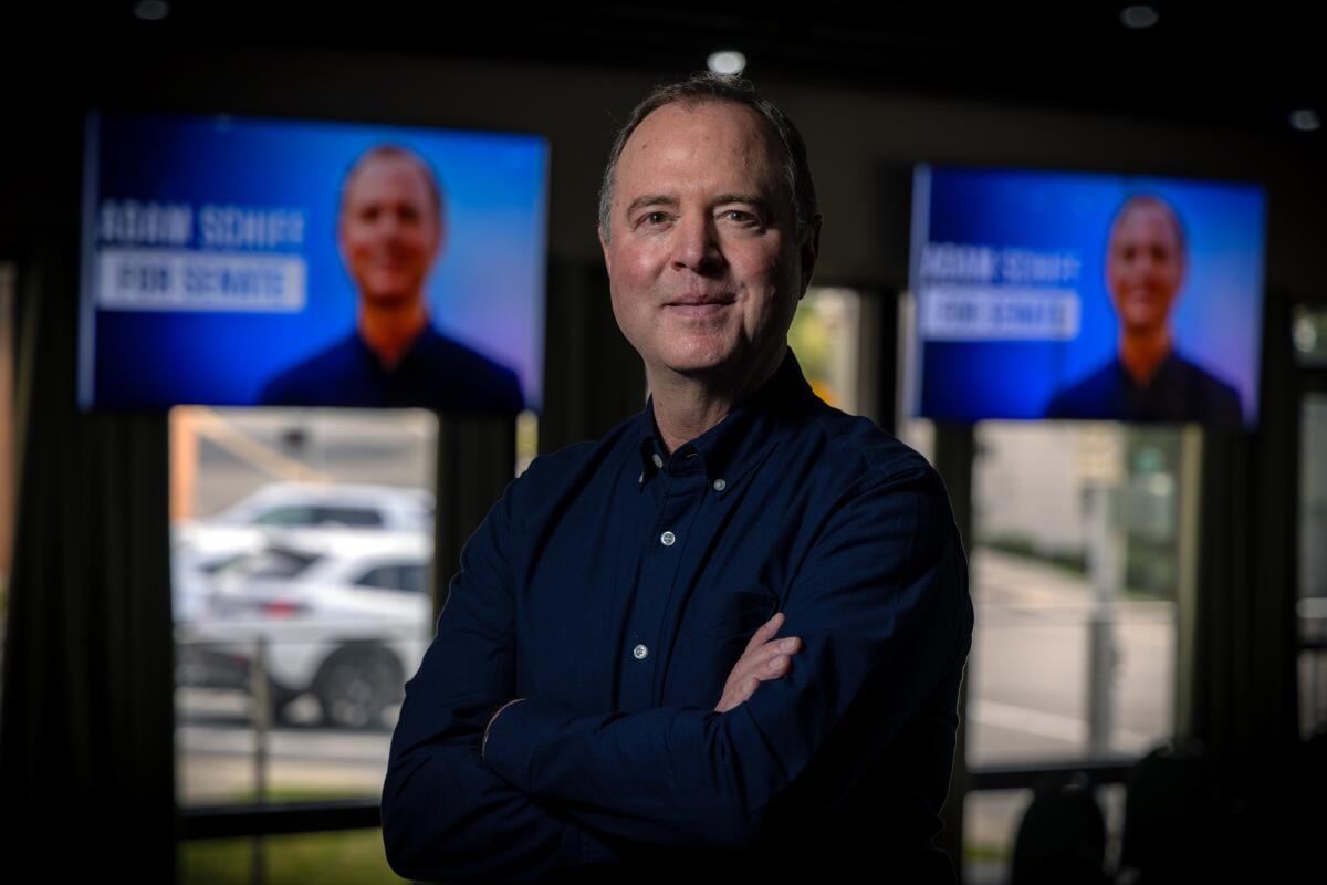 A man in a dark blue suit stands, smiling, with arms folded and images of him displayed in the background