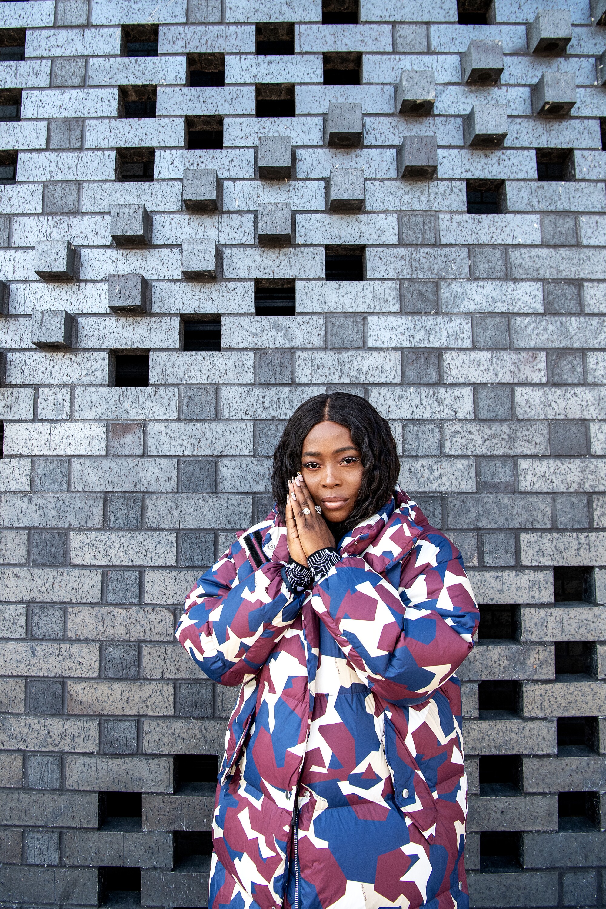 A woman in a colorful coat stands in front of a patterned wall.