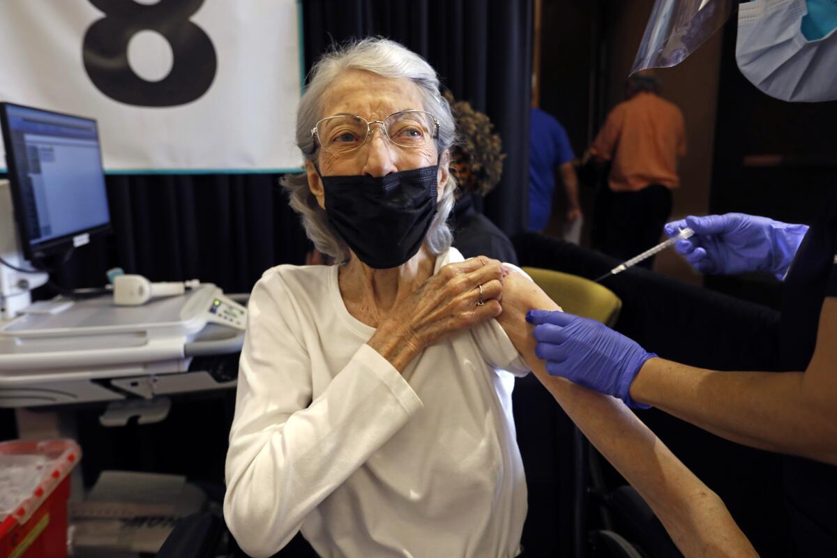 A woman wearing a white shirt and a mask is given a shot in the arm 