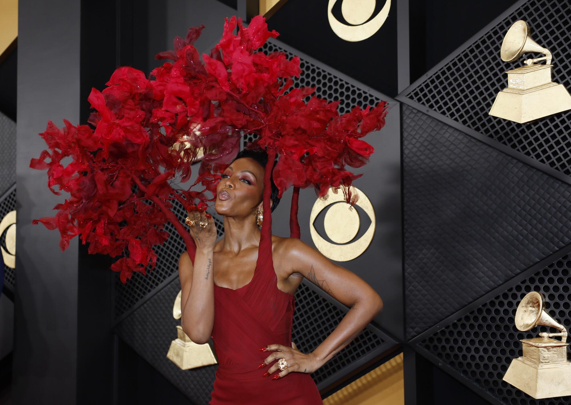 Dawn Richard blowing a kiss in a red dress that connects at the shoulders to an elaborate red floral headpiece