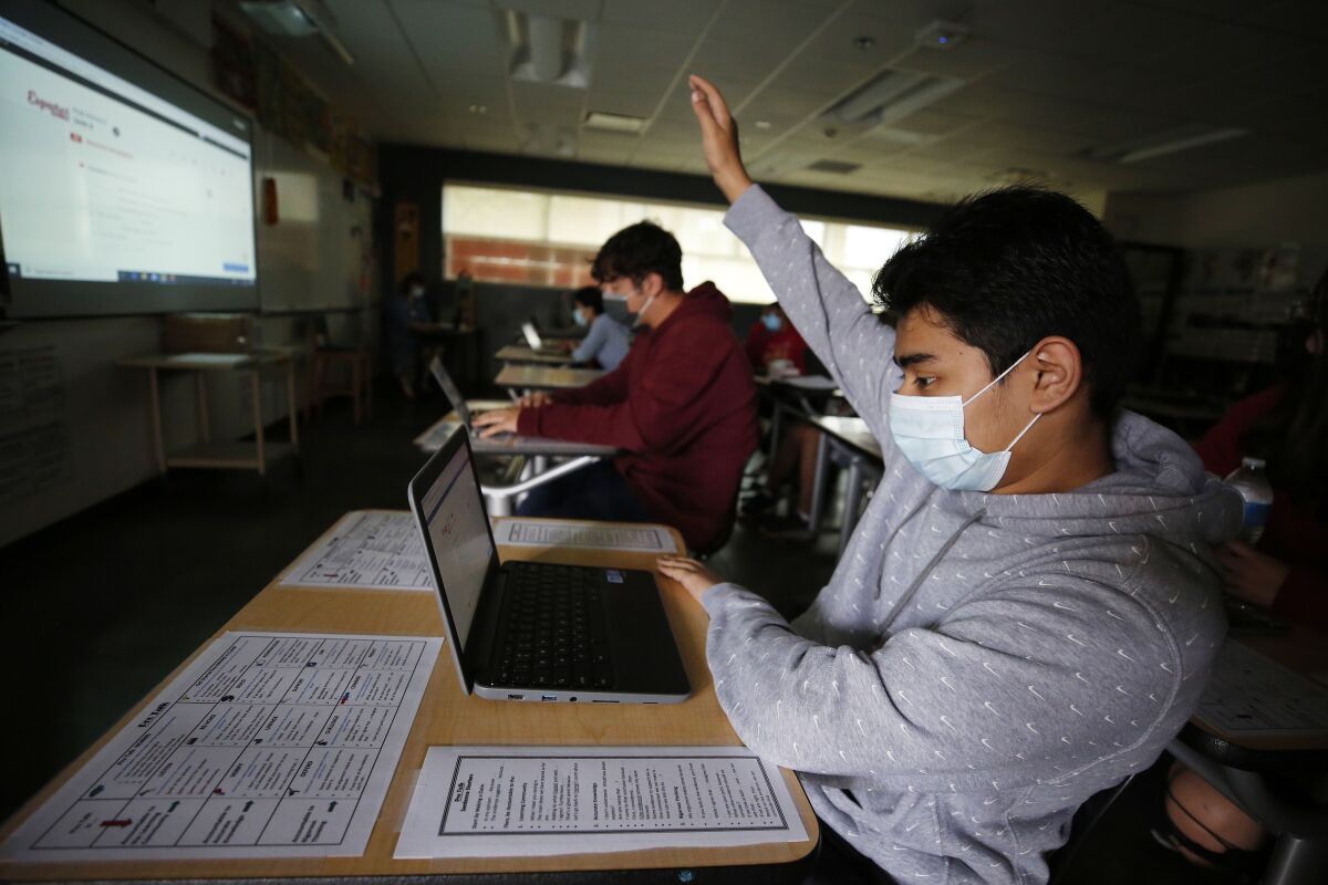 A masked student sitting at a desk with a laptop in a classroom raises his hand.