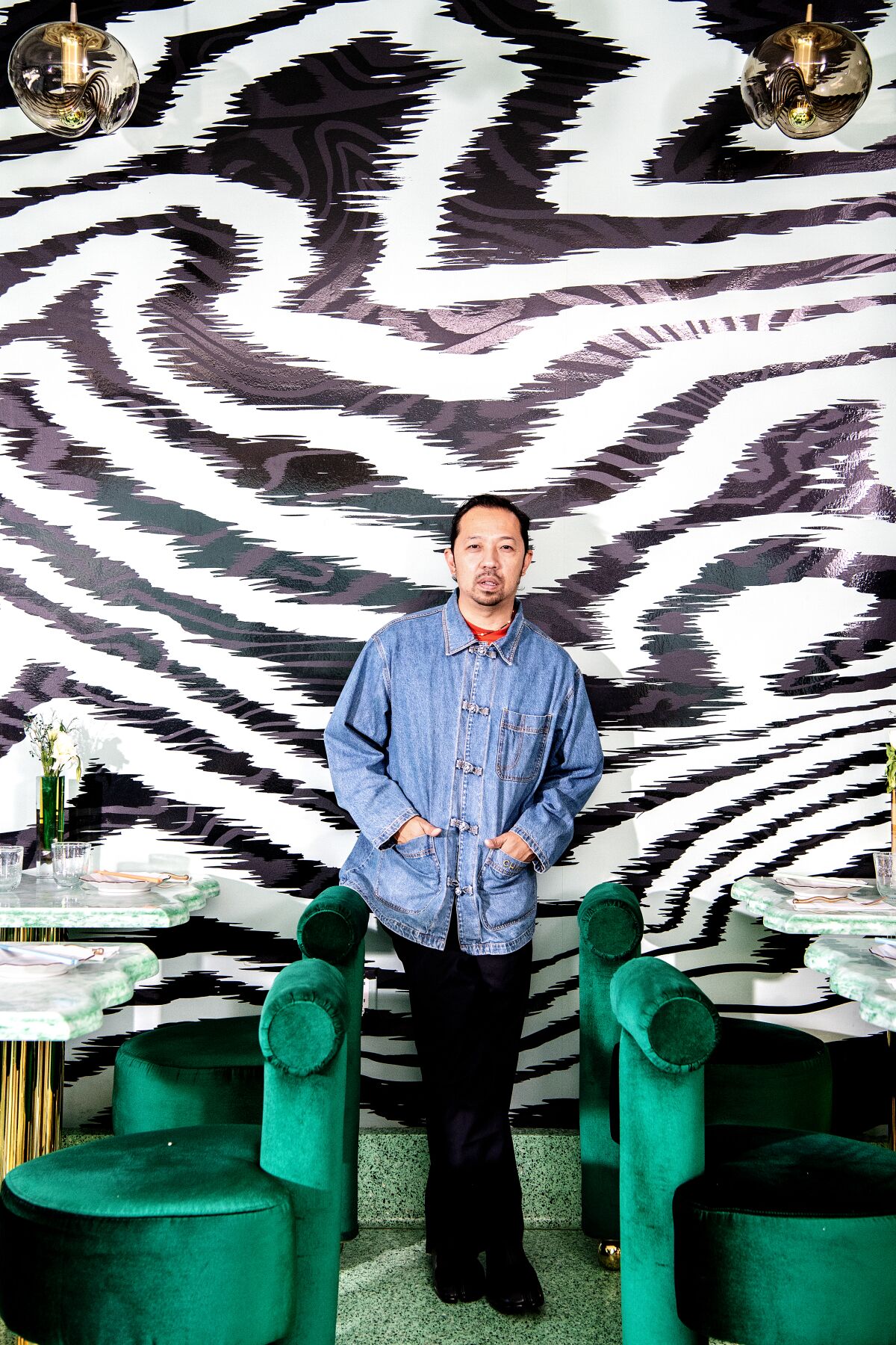 A man in a denim jacket stands between emerald green chairs against a zebra-print wall.