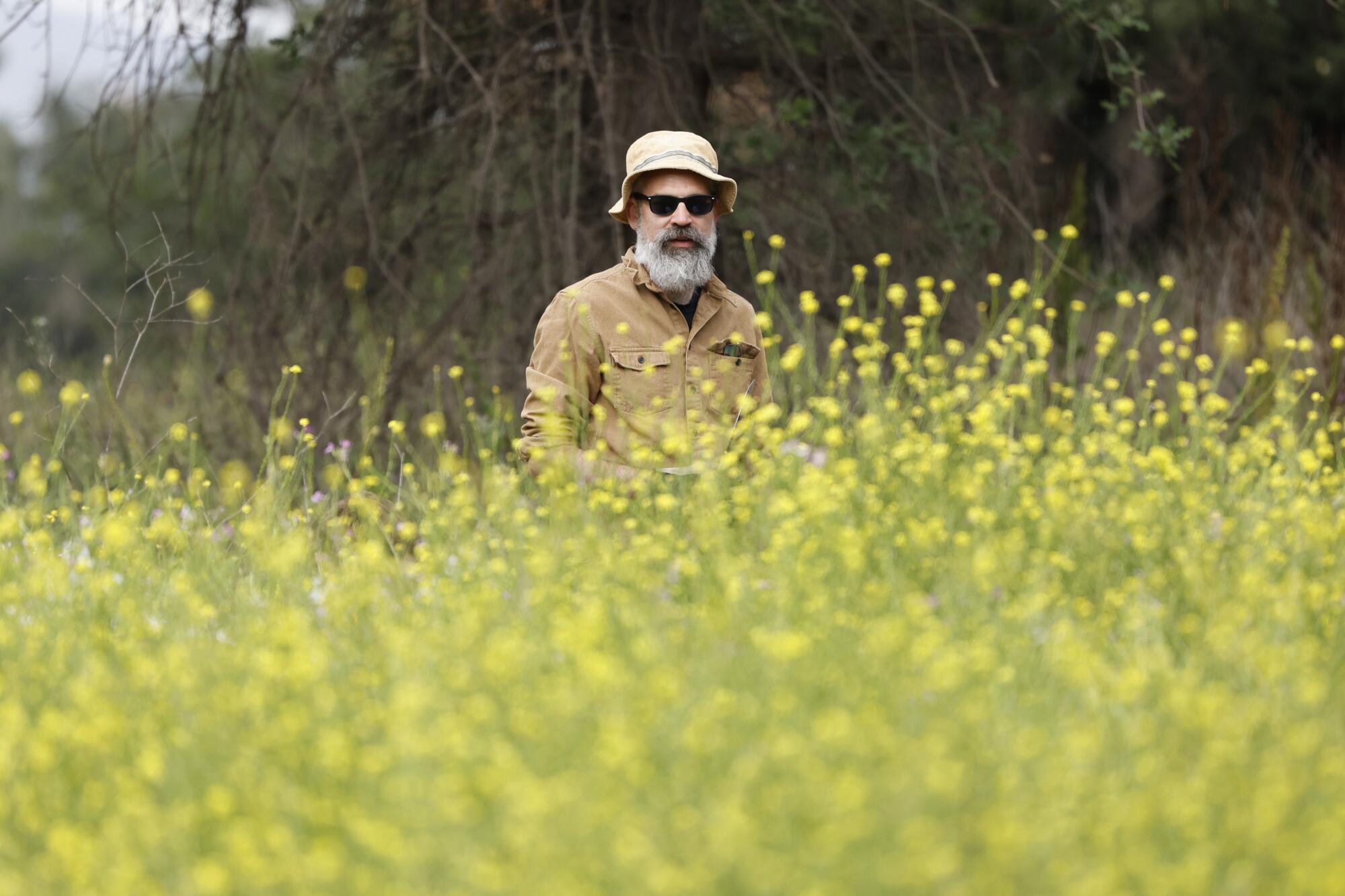A man stands behind a large number of plants with yellow flowers and brushes behind
