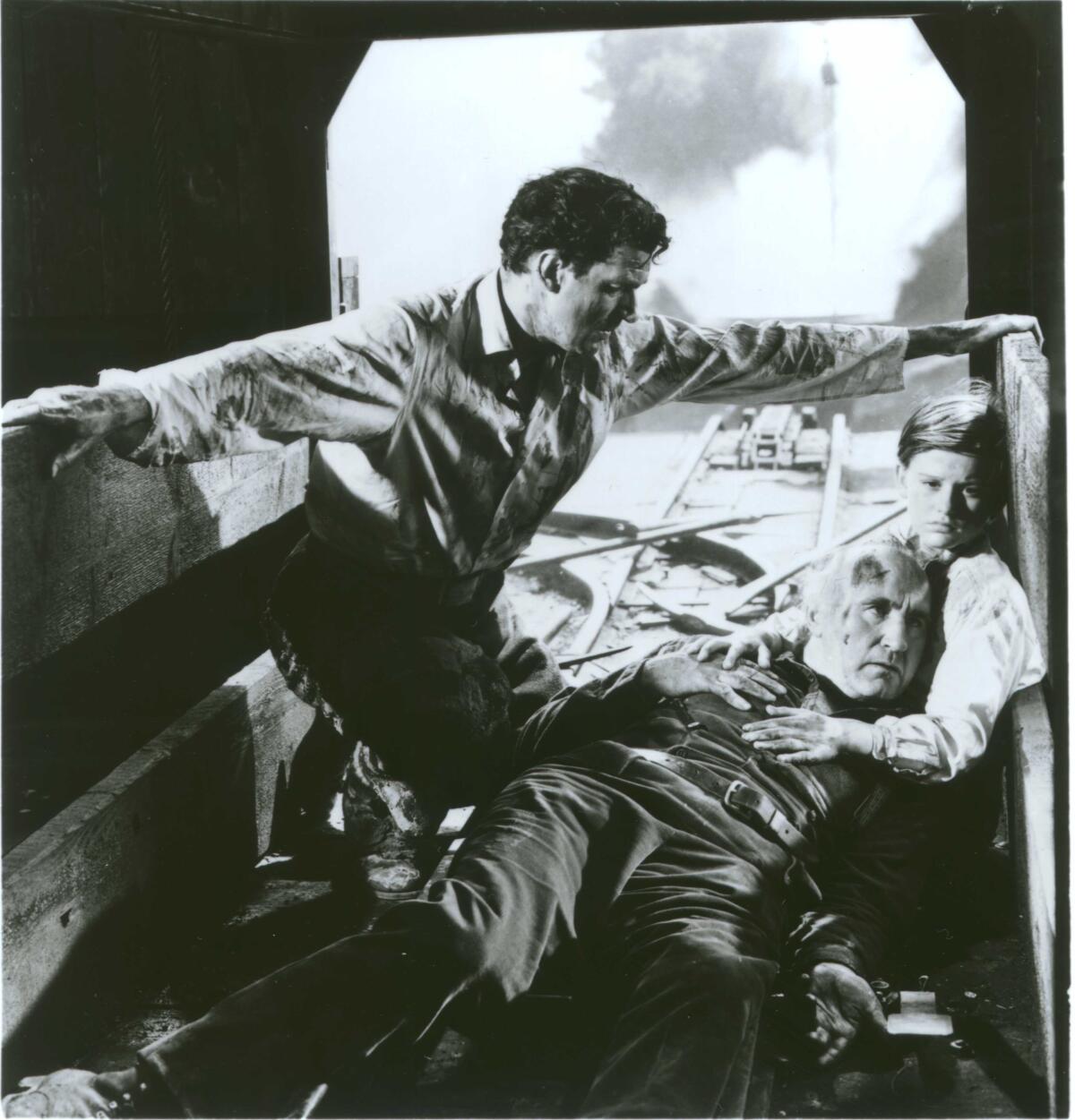 In a black-and-white movie still, a boy and a young man sit with an older man with a head injury in a coal mine.