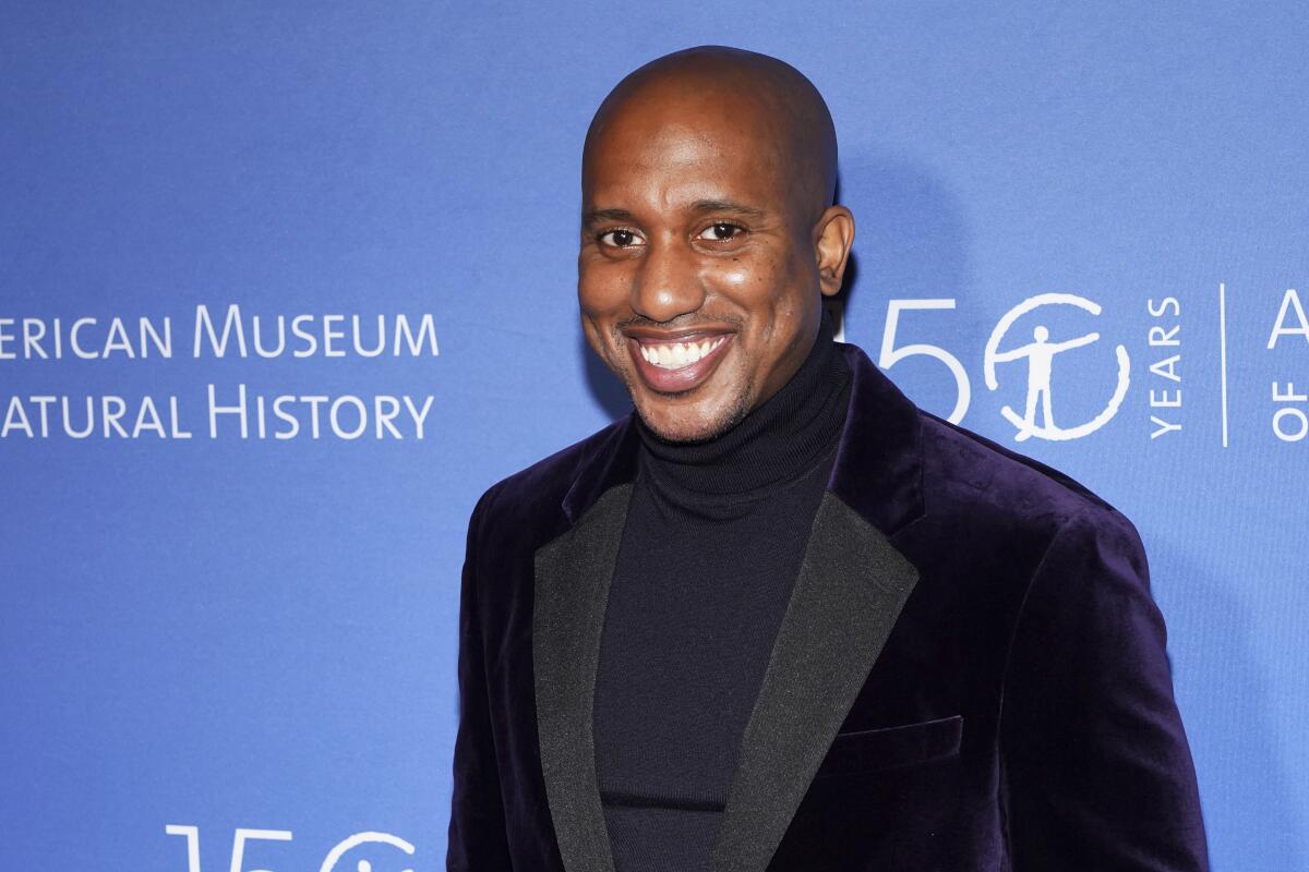 A bald man wearing a dark velvet suit and turtleneck smiles while standing in front of a blue backdrop