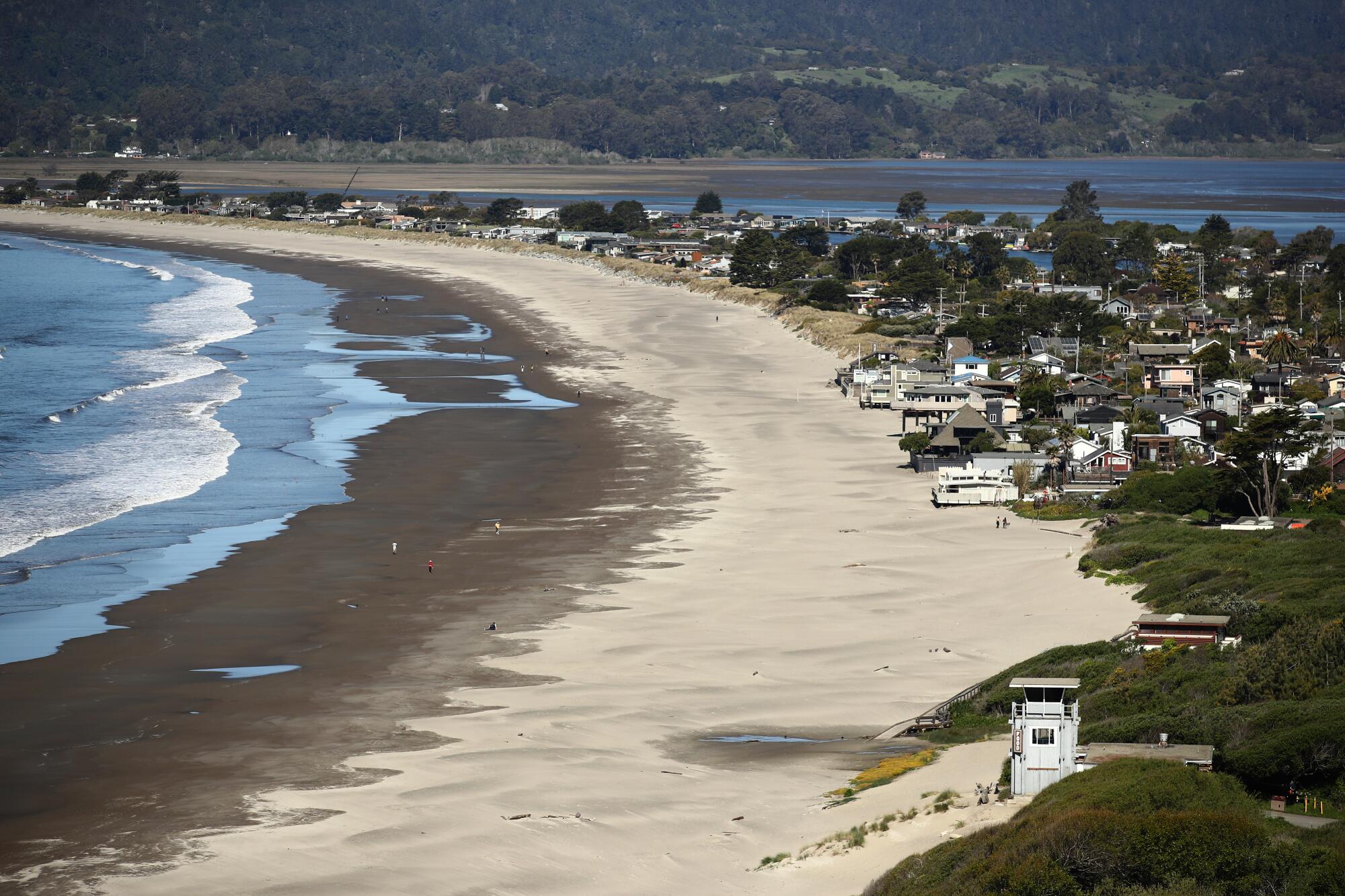 Homes lining a curving sandy shoreline on the ocean, with another body of water and green hills in the background
