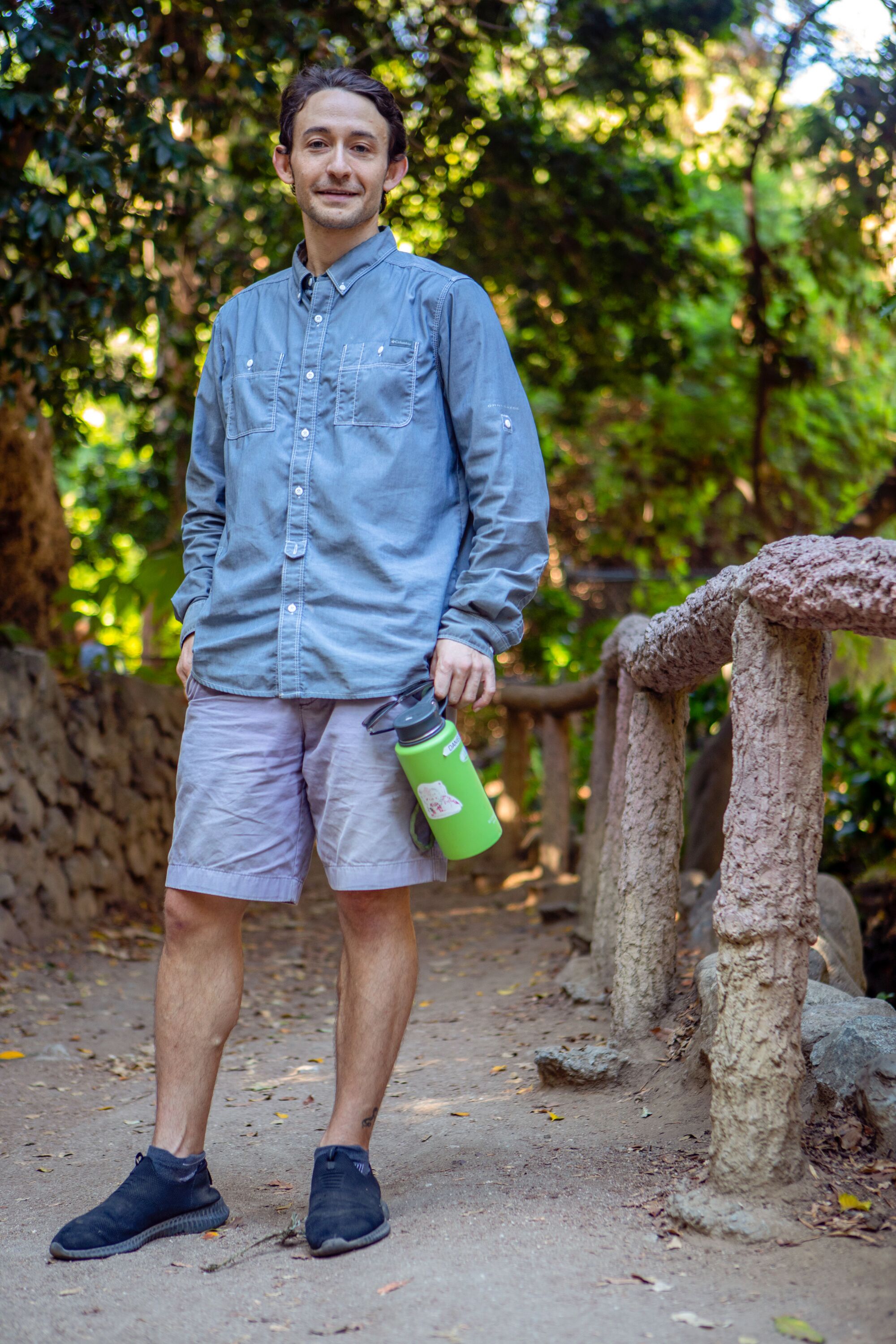 Şortlu ve yürüyüş tişörtlü bir adam dışarıda duruyor ve elinde yeşil bir su şişesi tutuyor.