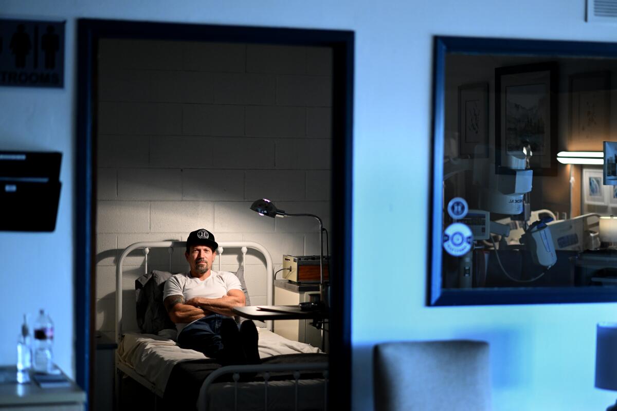 A man in a black hat sits on a hospital bed.