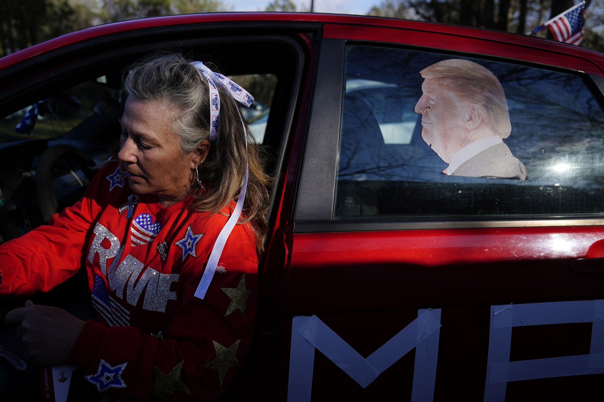 A woman with a red shirt with the word "Trump" in glitter gets out of a car, an image of Trump on one window.