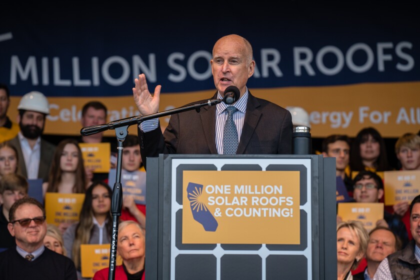 A man speaks into a microphone at a  lectern with the sign "One million solar roofs & counting."
