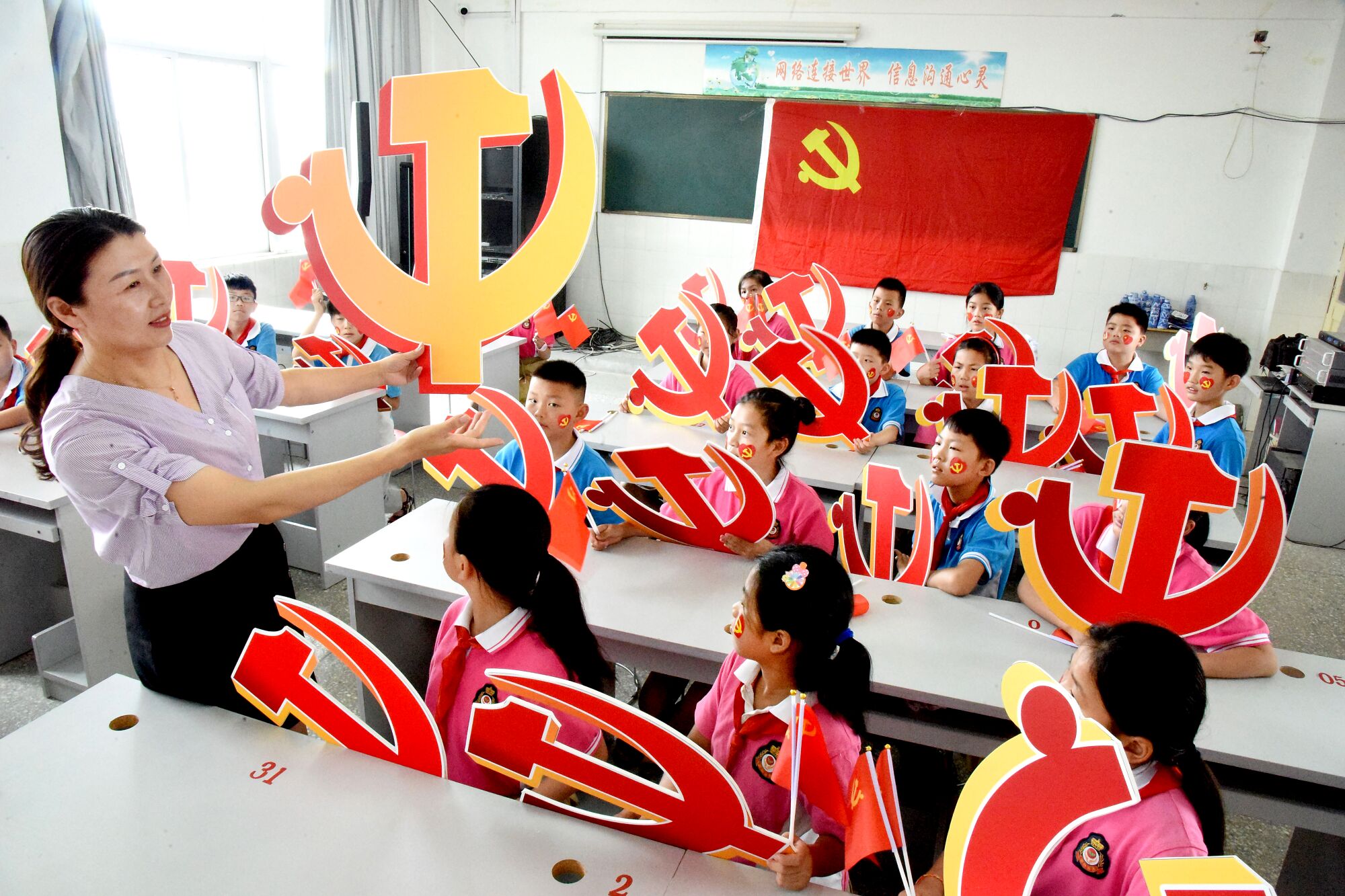 A woman holds a hammer and sickle emblem as children watch, holding their own red-and-gold emblems 