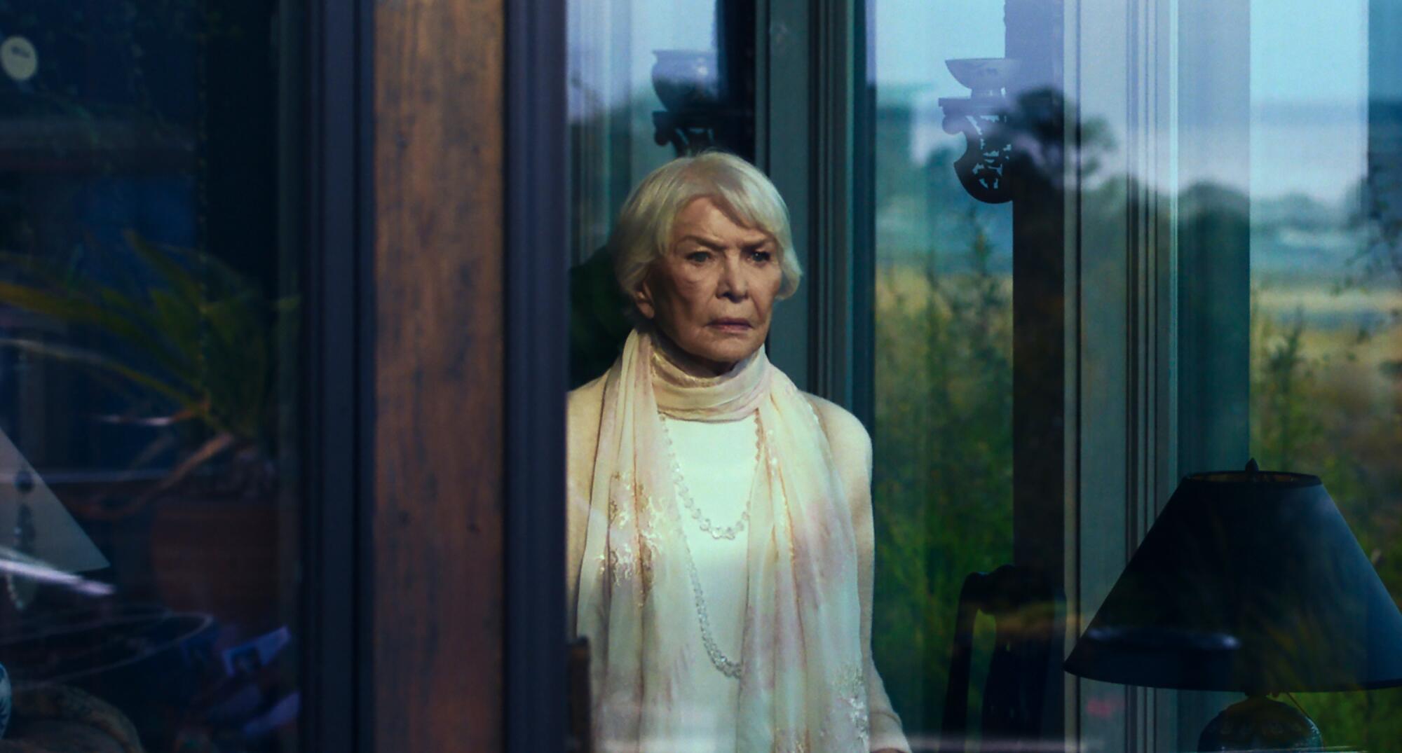 A graying, fearful woman stands at a window.