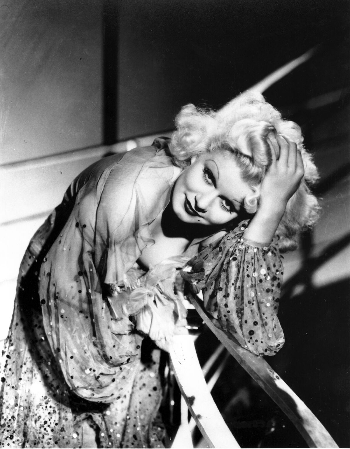 A woman with white blond hair and dramatic makeup and wearing a sparkling gown leans against a railing.