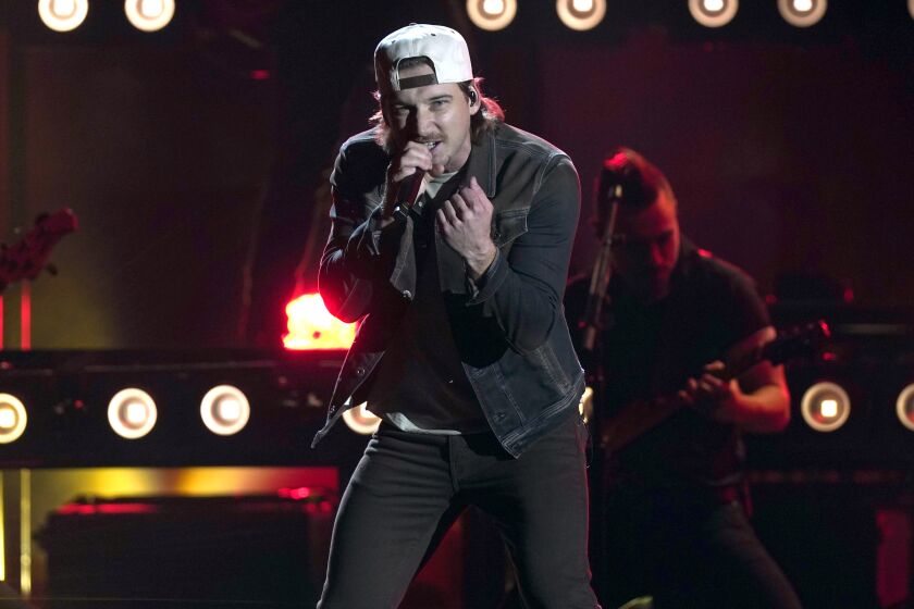 A man wearing a black outfit and backward white hat while singing into a microphone and balling his hand in a fist onstage