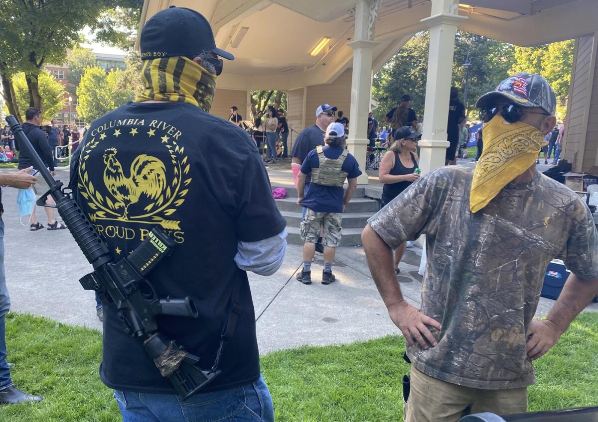 A man in a Proud Boys T-shirt carries a rifle and talks with another man at an outdoor gathering in September.