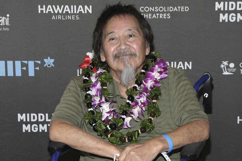 A man in a wheelchair with dark hair and a gray beard wearing a green shirt and multiple leis around his neck