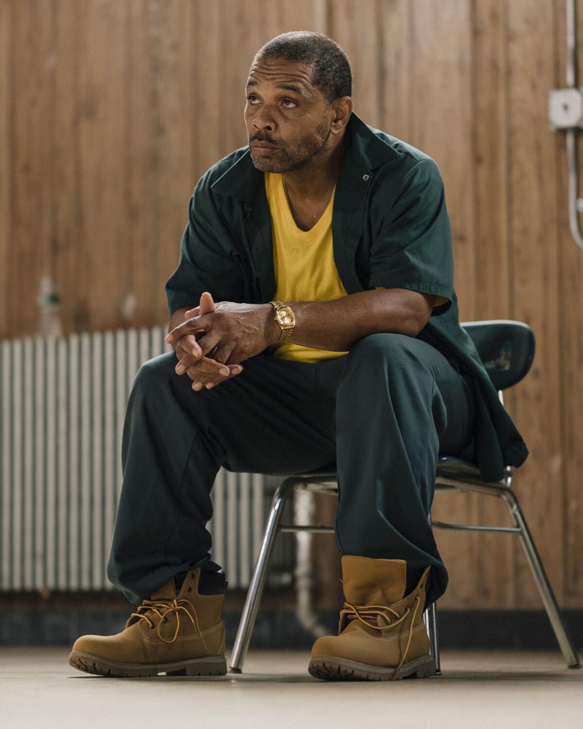 A man in boots and a yellow T-shirt sits on a chair.