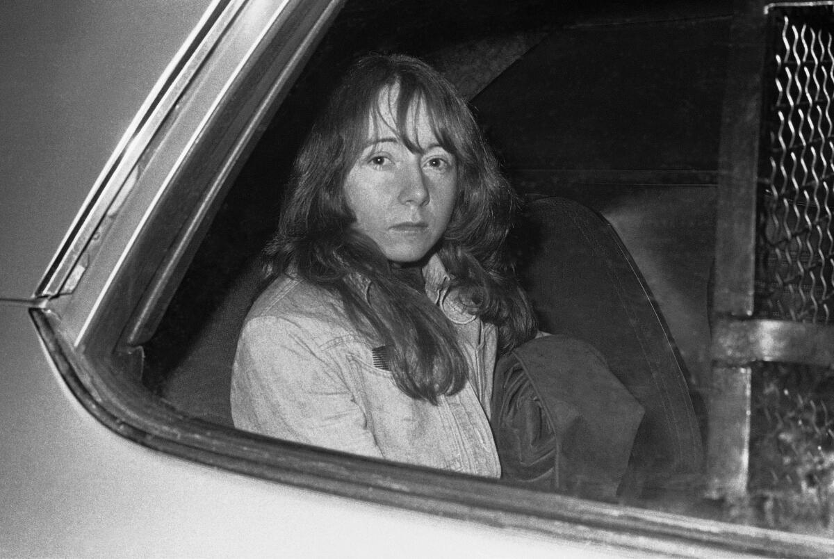 A young woman with long hair and bangs sits unsmiling in the back seat of a car.