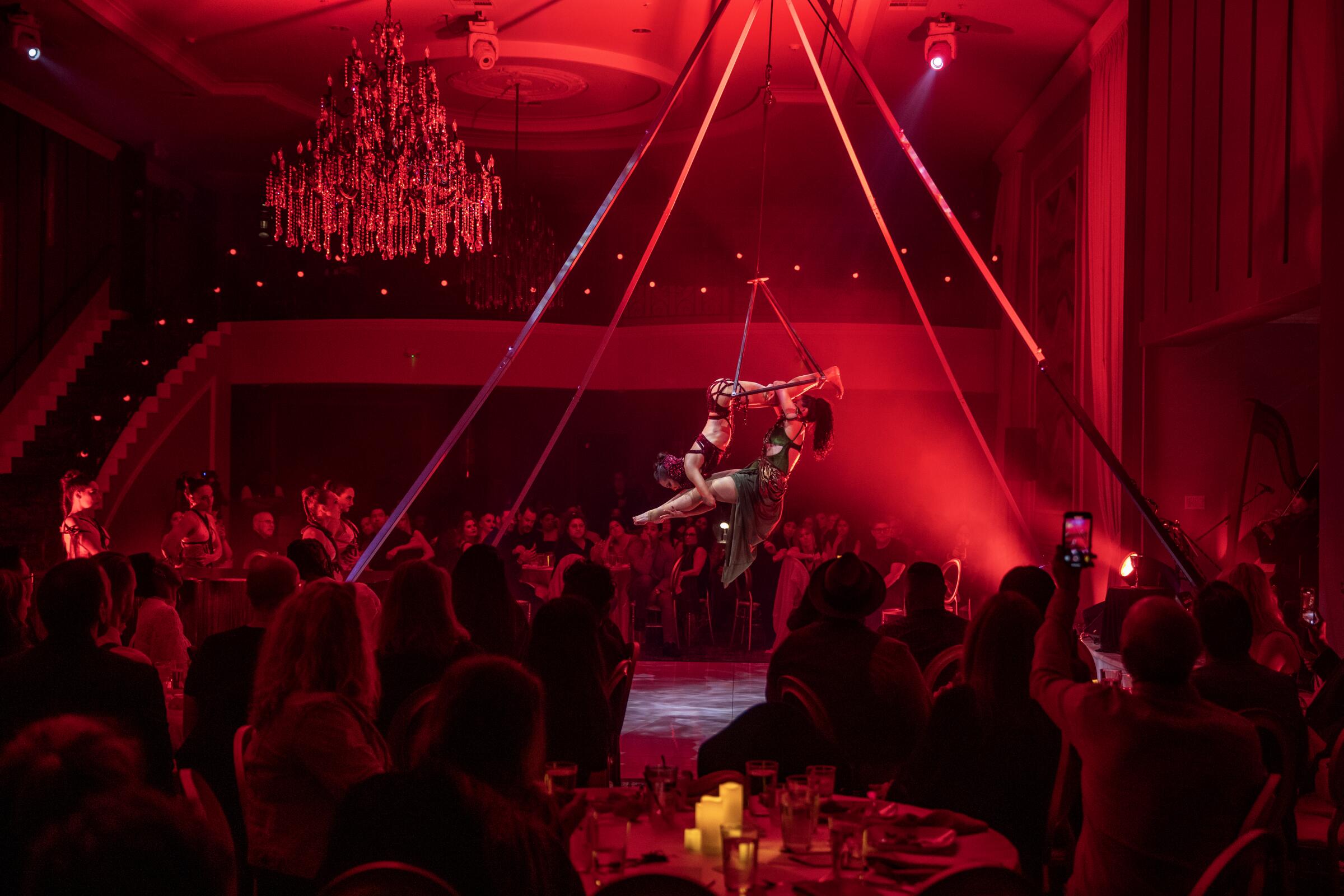 Two women in black perform an aerialist show in a red-lighted theater with an audience.