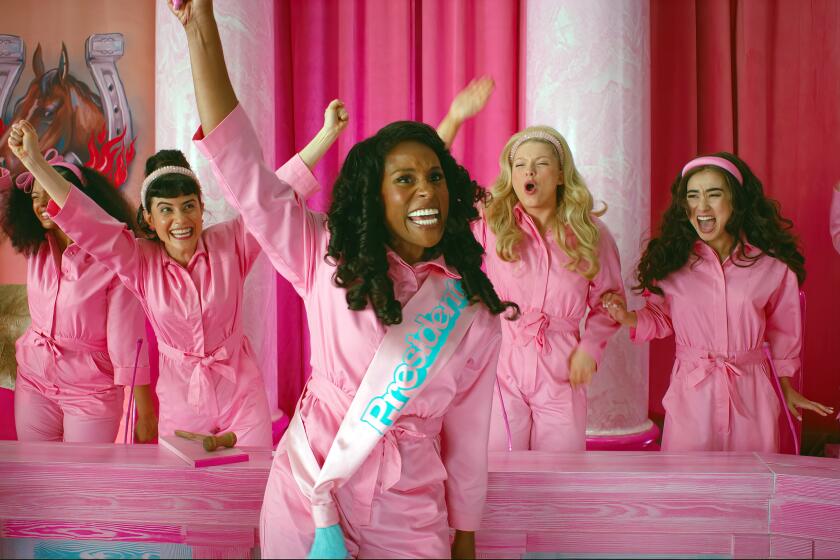 Issa Rae in a pink jumpsuit and a pink sash holding up her right hand surrounded by women wearing similar outfits