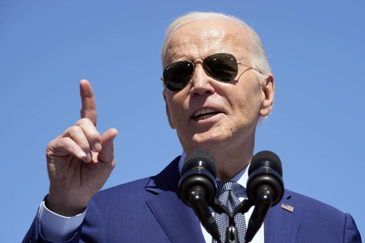 Biden, seen from the shoulders up in a blue suit and tie, points upward as he speaks into two microphones under a clear sky