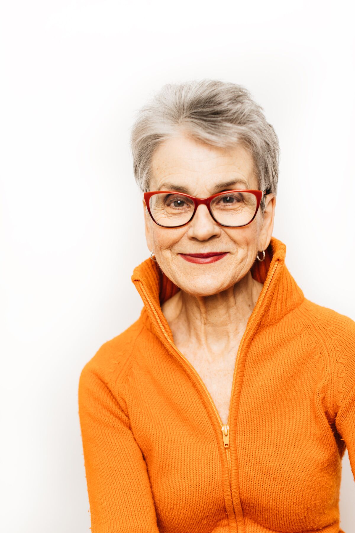 Author Frances Moore Lappé wears an orange zip-up sweater and red glasses, smiling at the camera.