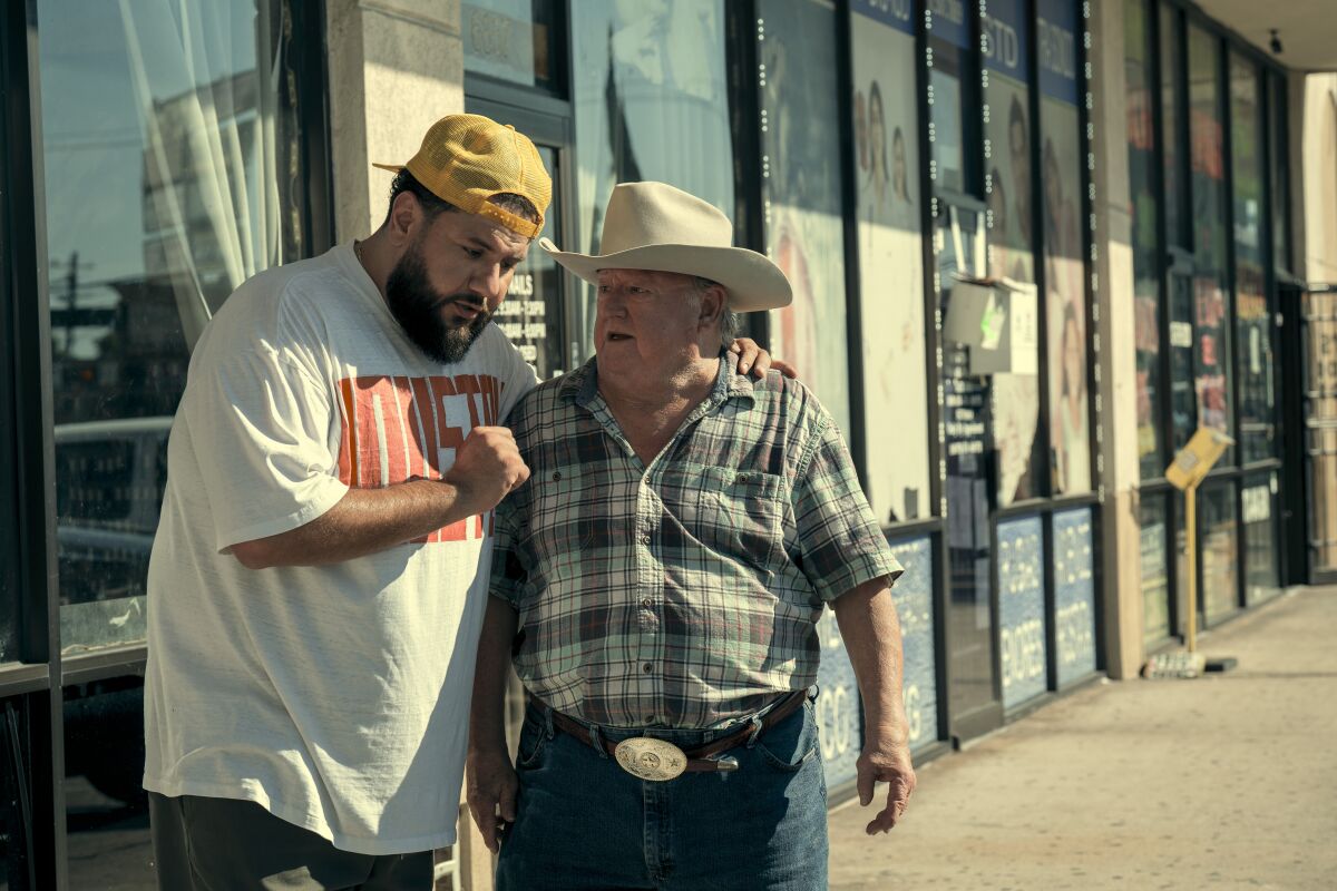A man in a backward baseball cap with his arm around a man in a cowboy hat on the sidewalk outside stores.