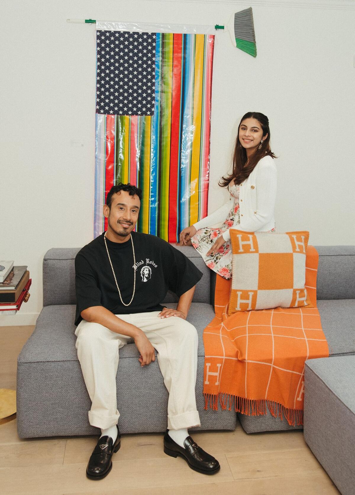 A man sits on a couch next to a woman sitting on the back of the couch in front of a rainbow American flag.
