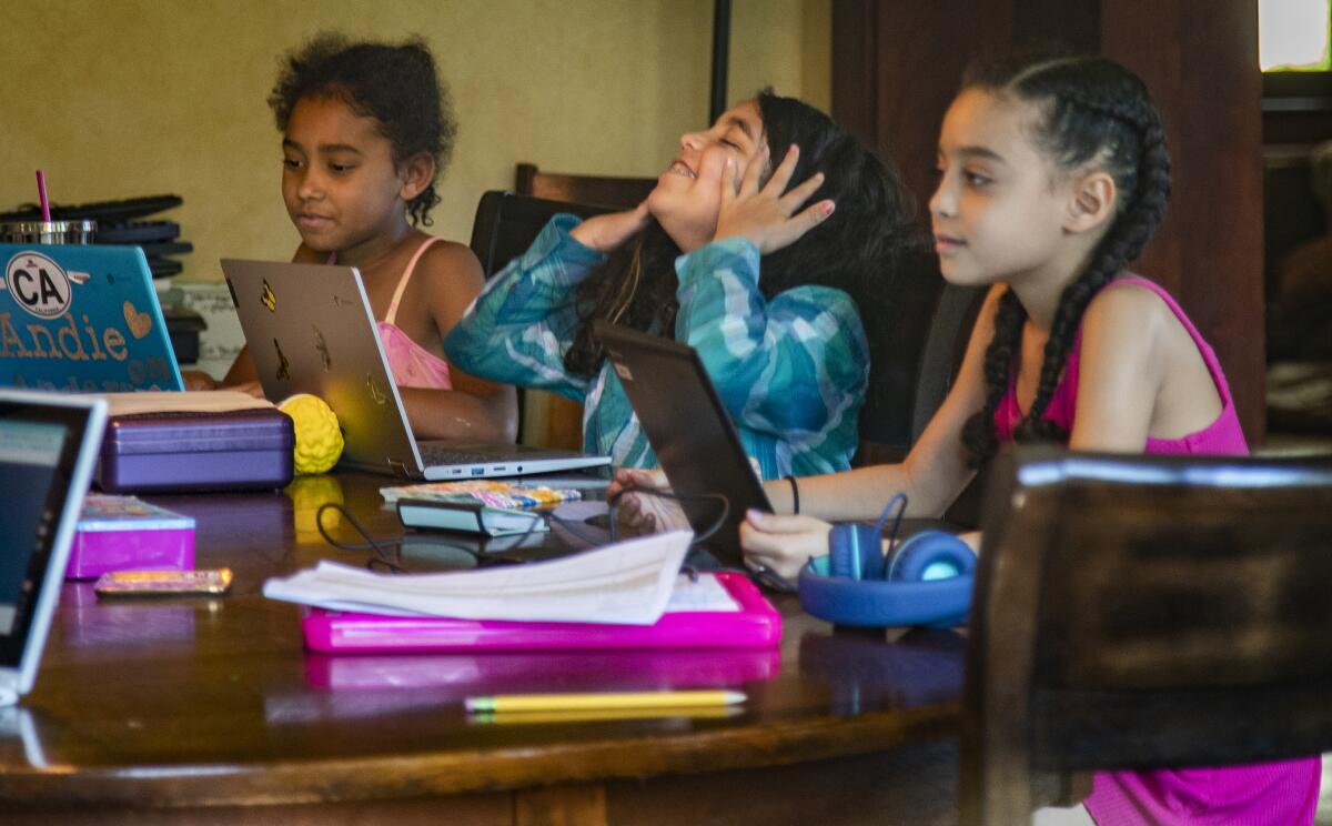 Three girls sit at a dining room table with laptops doing homework.