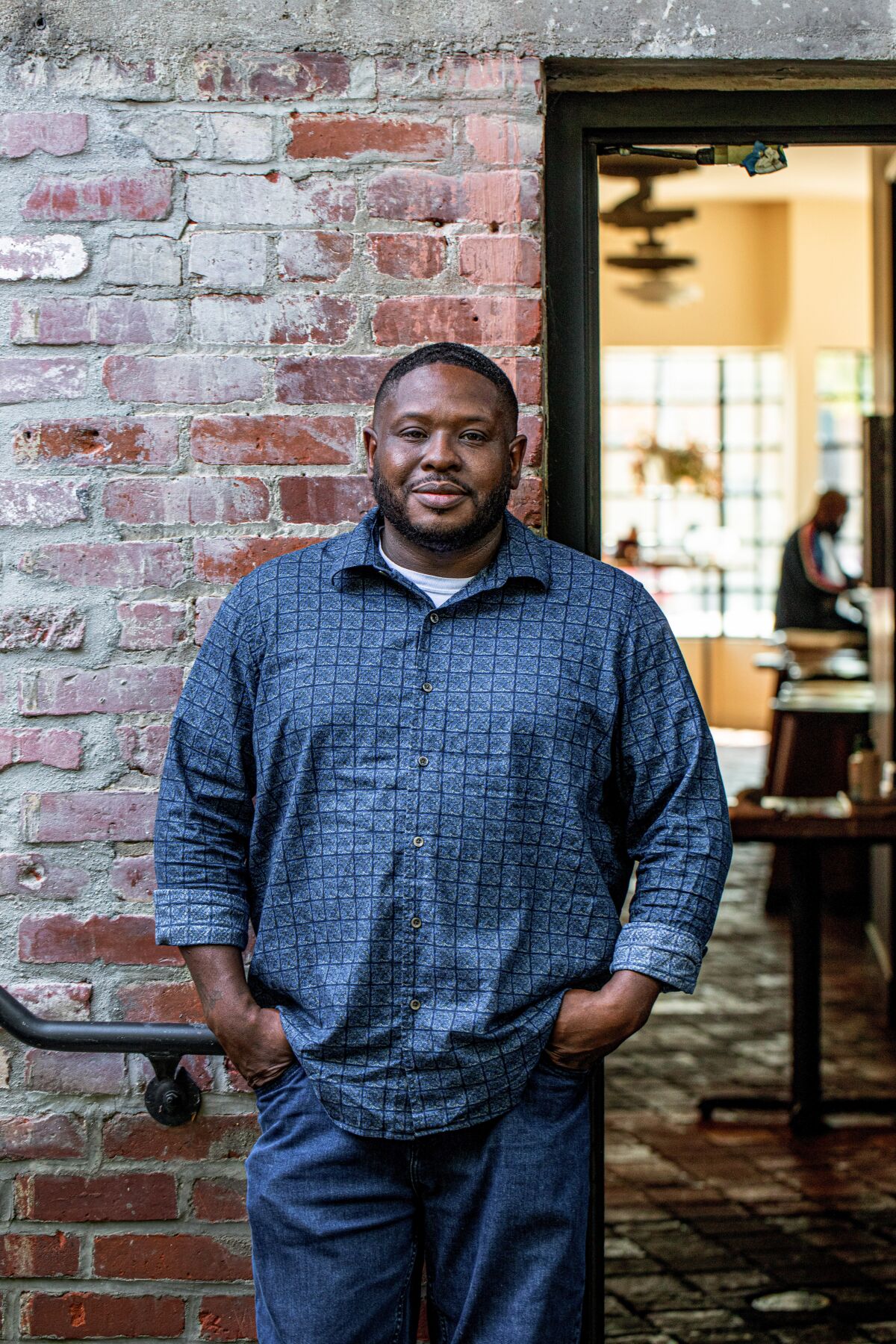 A man in a blue button-down in jeans stands in front of a brick wall