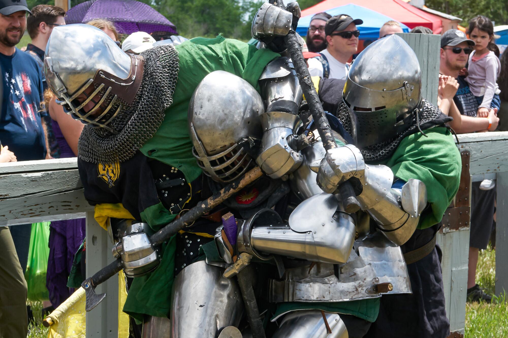 People in armor grapple, holding medieval-style weapons, as spectators watch on the other side of a wooden fence.
