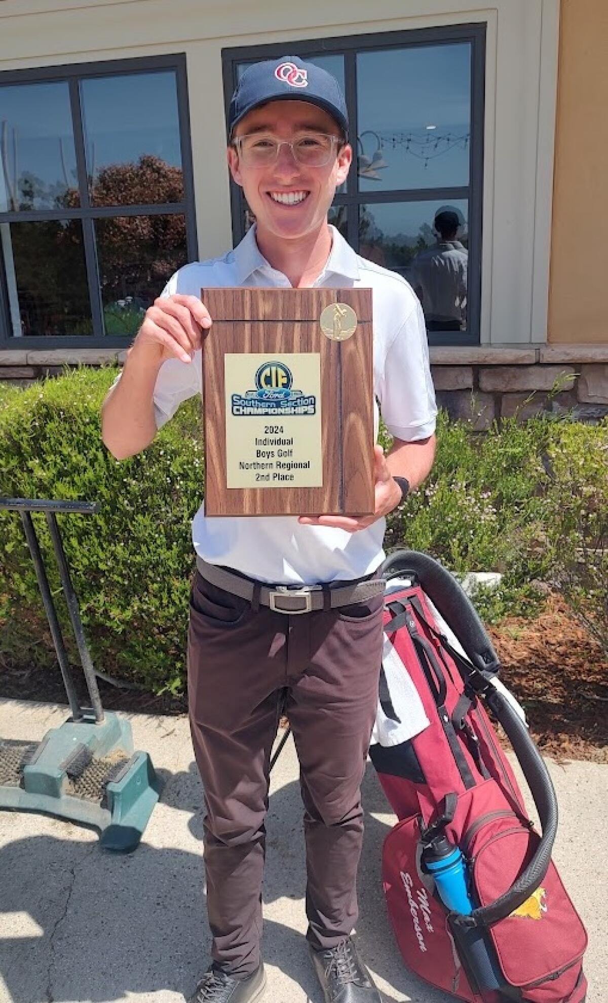 A smiling Max Emberson holding a plaque with a golf bag at his feet 