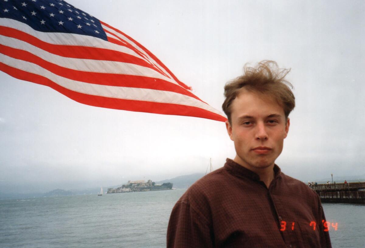 A young man stands with a waving American flag and a body of water behind him.