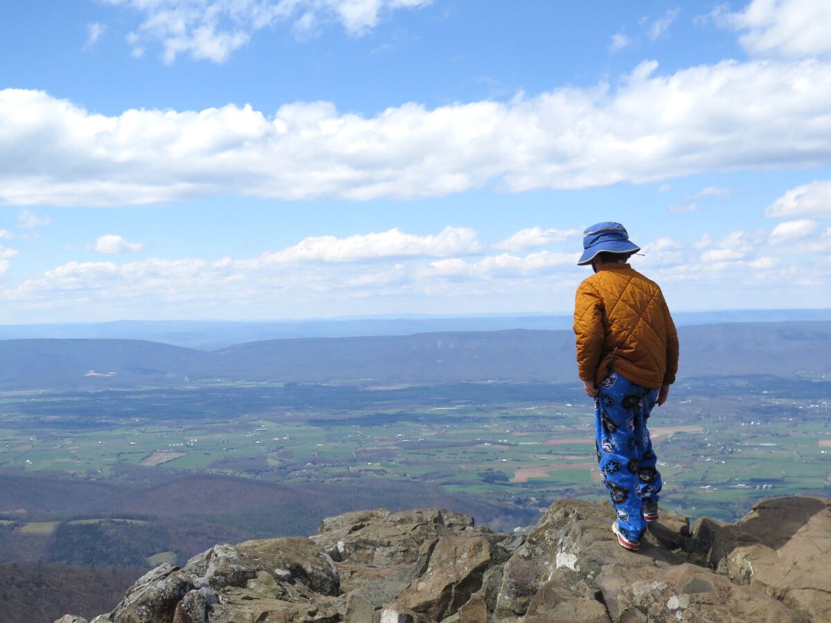 A boy in pajama pants, a hat and jacket stands on a rock outcropping overlooking a valley.