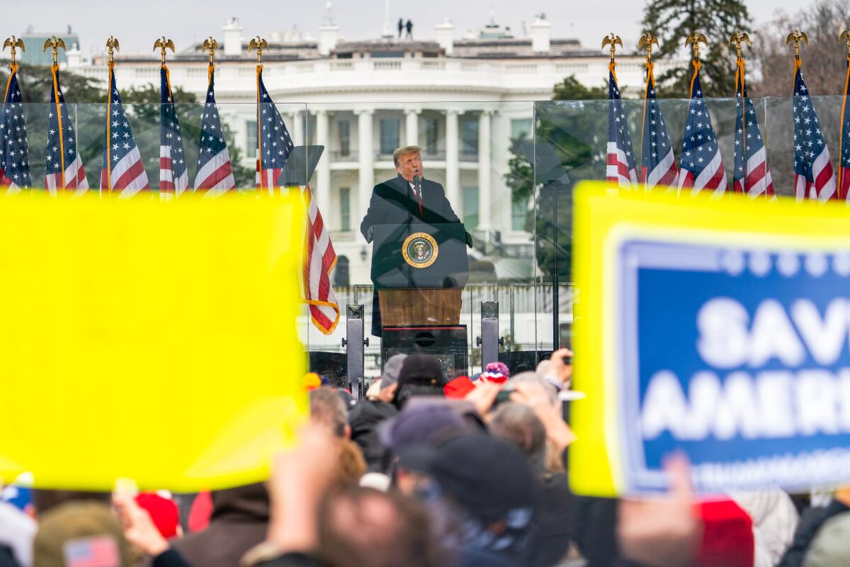 A man in a dark overcoat, flanked by U.S. flags, speaks at a lectern outside the White House before a crowd