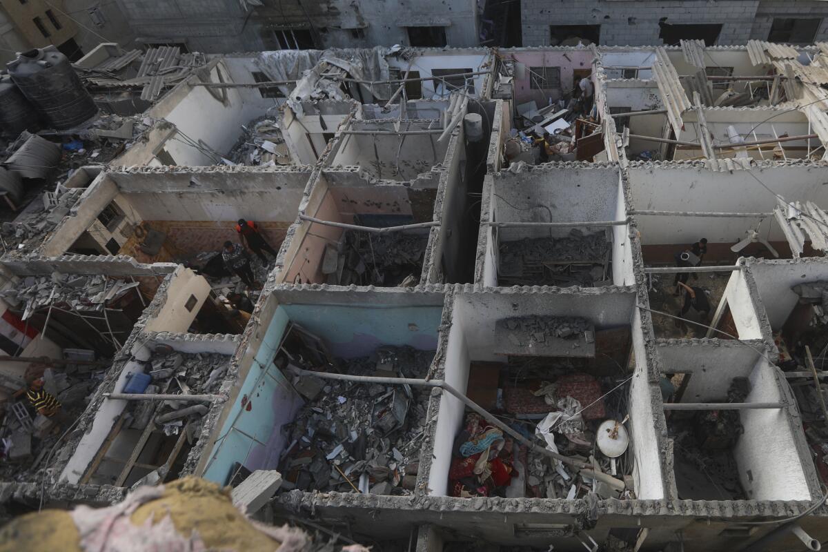 A bird's-eye view of exposed rooms in buildings missing roofs and people inside, amid rubble 