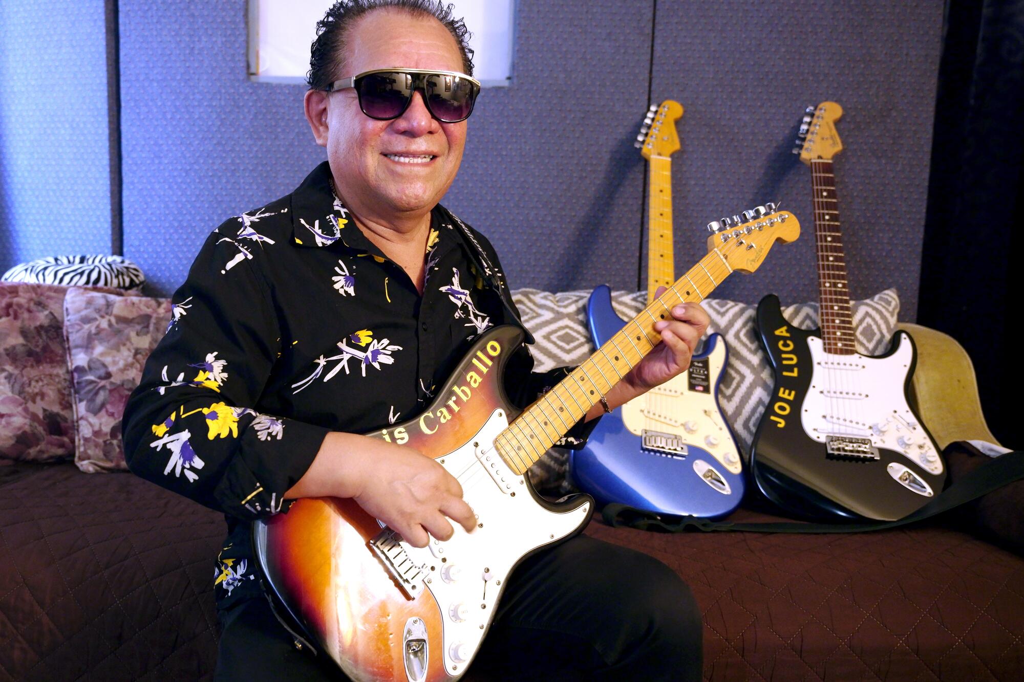 A man in sunglasses plays the guitar in front of a couch with two guitars on it 