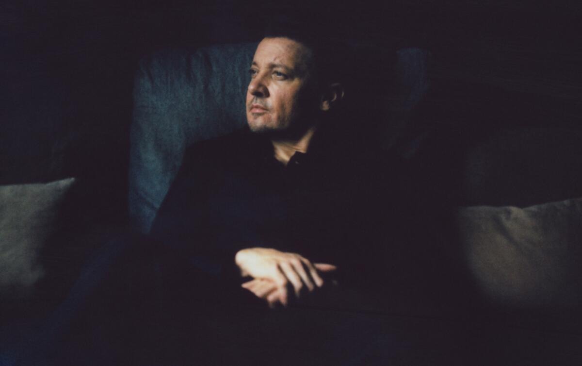 Jeremy Renner sits, looking off into the distance, wearing a black collared shirt.  