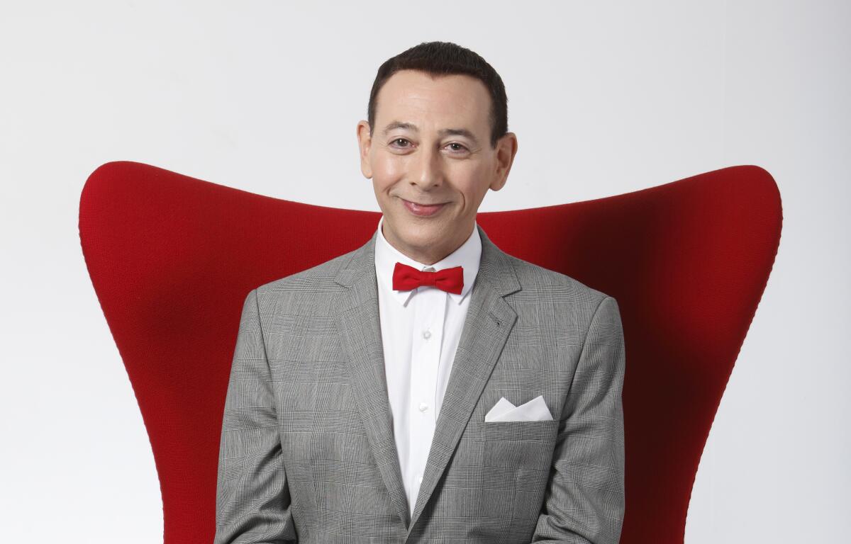 A smiling Paul Reubens, wearing a gray suit and red bowtie, sits in a red chair. 