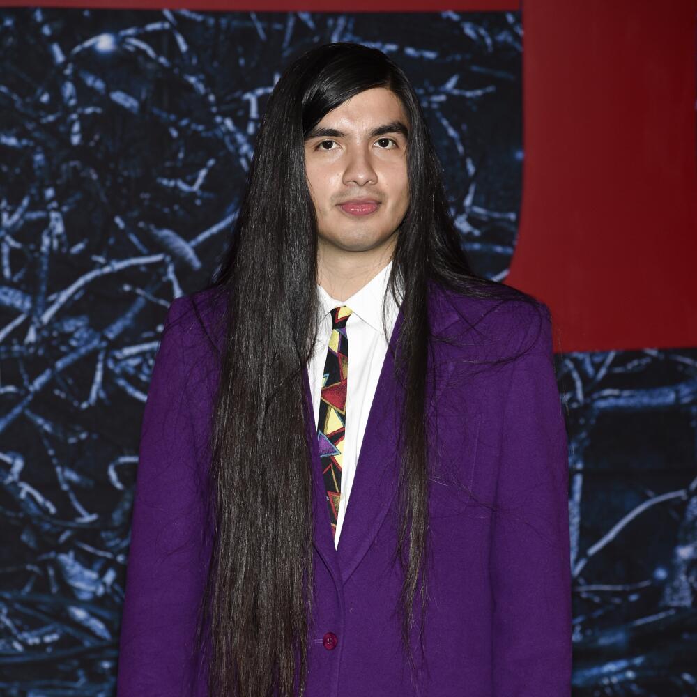 A man with long brown hair, wearing a purple blazer, poses for a photo
