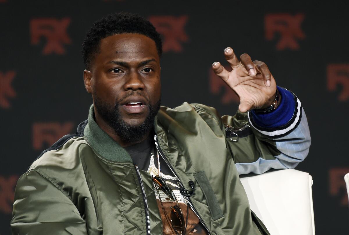 Kevin Hart in a shiny olive jacket raising his hand