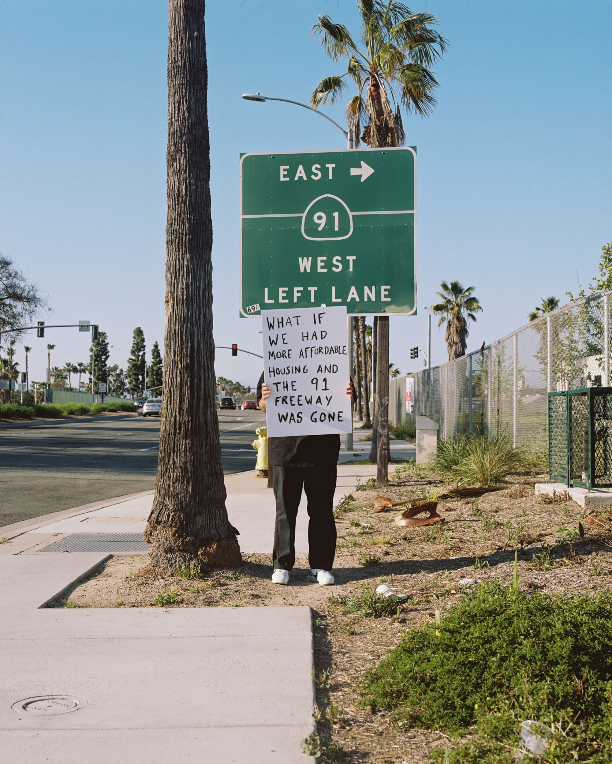 A man stands beneath the East 91 highway sign and holds up a sign reading, "What if we had more affordable housing"