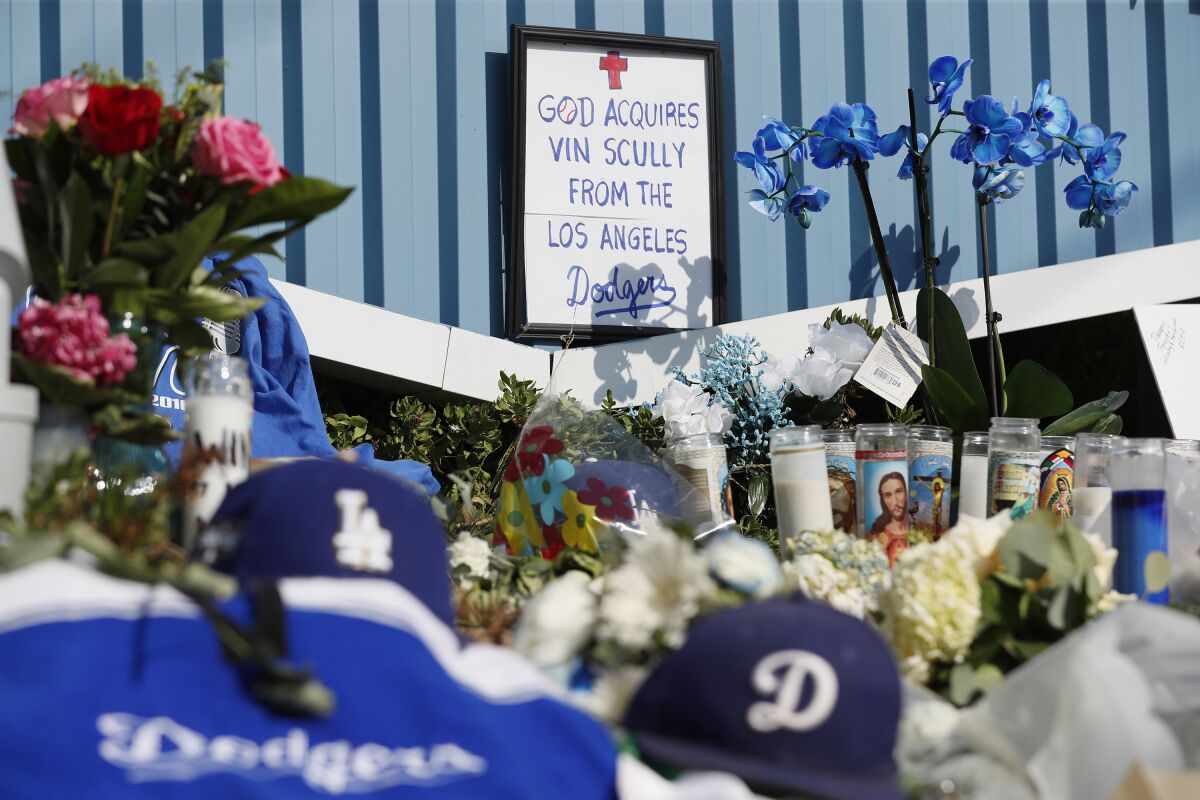 a pile of flowers, candles and memorabilia below a sign that reads: 'god acquires vin scully from the los angeles dodgers'