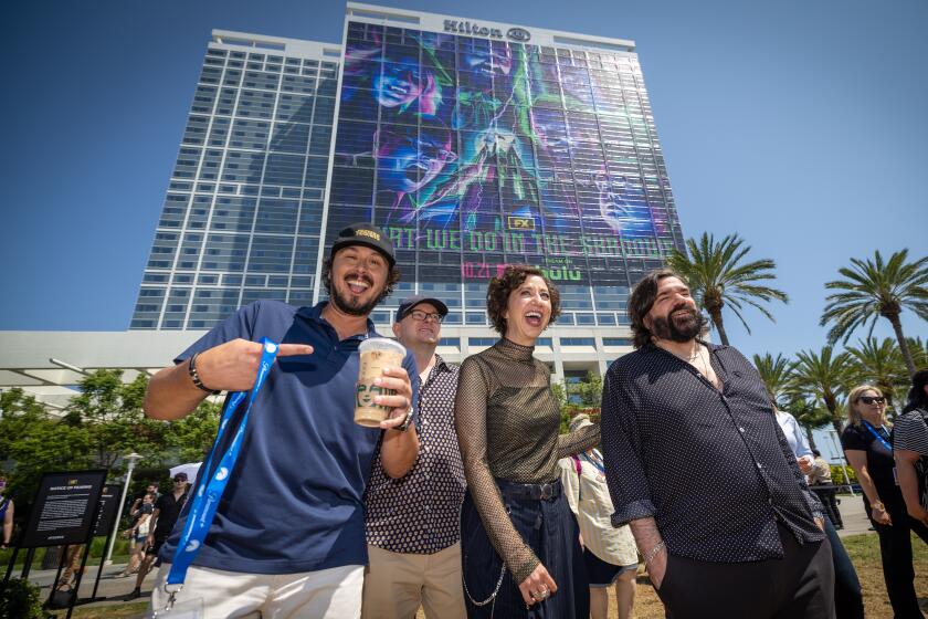 San Diego, CA - July 25: From left: Kyle Newacheck, EP/Director, actor Mark Proksch, who plays "Colin Robinson," actor Kristen Schaal, as "the guide", actor/writer/composer Matt Berry, as "Laszlo" of the FX comedy "What We Do in the Shadows" arrive at the FX Lawn to tour the "Shadows" activation & complete the fan interaction with a view of a giant advertisement that covers the front of the Hilton Bayfront in the background at San Diego Comic-Con Thursday, July 25, 2024 in San Diego. The series is ending with its forthcoming sixth season. (Allen J. Schaben / Los Angeles Times)