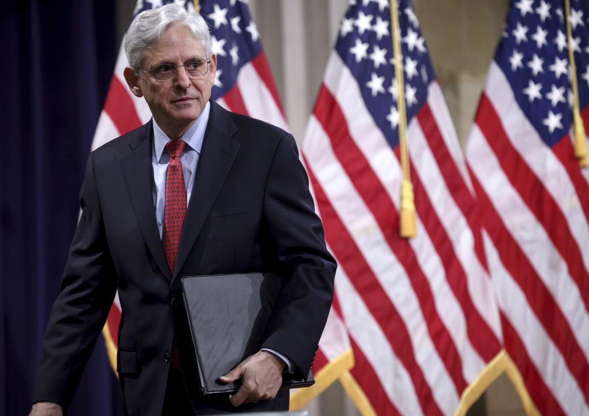 A man in glasses and a suit holds a folder against a backdrop of U.S. flags 