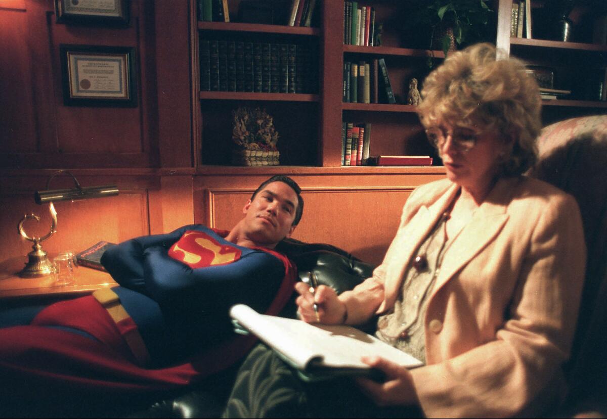 Superman reclines on a couch as he speaks to a woman in a beige blazer and top who is holding a notepad and pen.